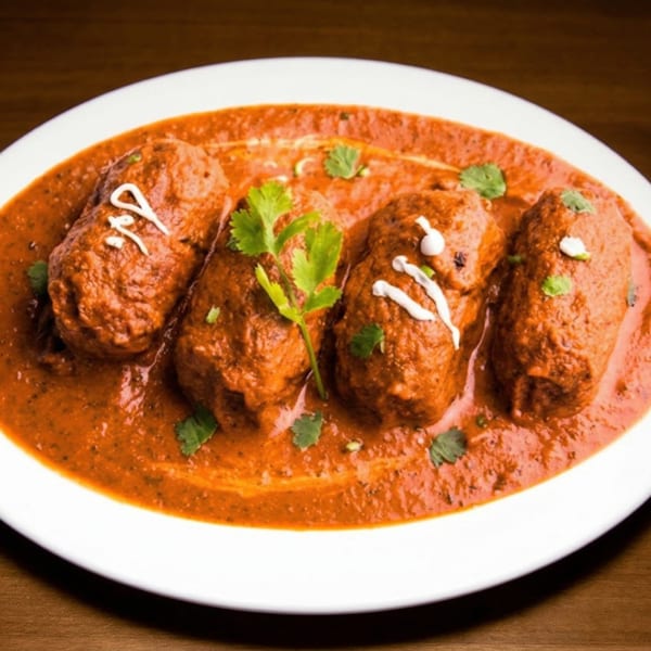 Four Indian kofta dumplings in a rich red curry sauce, garnished with fresh cilantro and cream swirls.