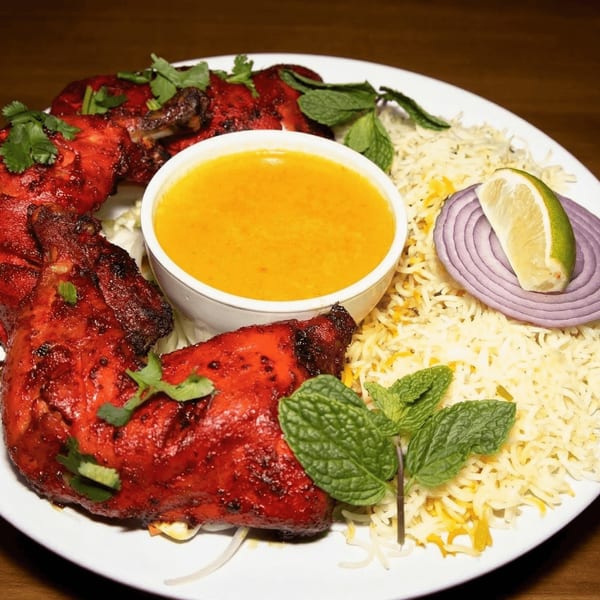Tandoori chicken with rice, a bowl of yellow sauce, mint, red onion, and lime on a white plate.